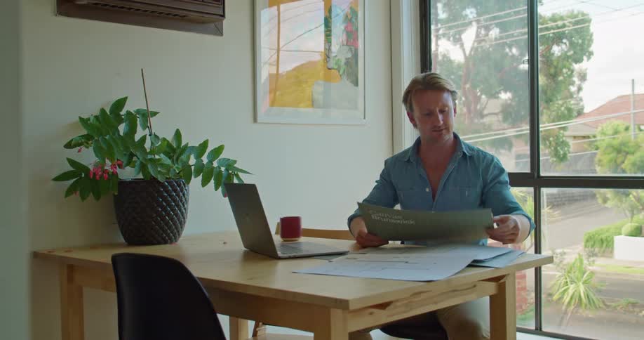 A man picks up and looks at different plans while sitting at his desk with a laptop