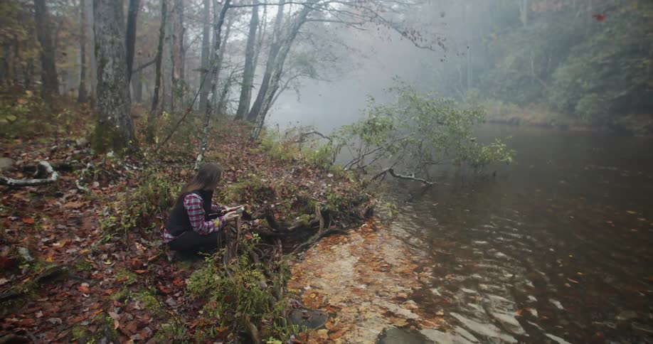 Woman sits on bank in woods near floating fallen leaves with mobile phone