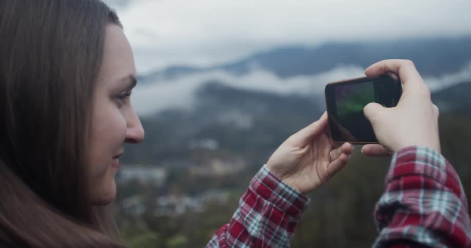 Smiling woman standing outside of small town takes photograph of landscape using her mobile phone