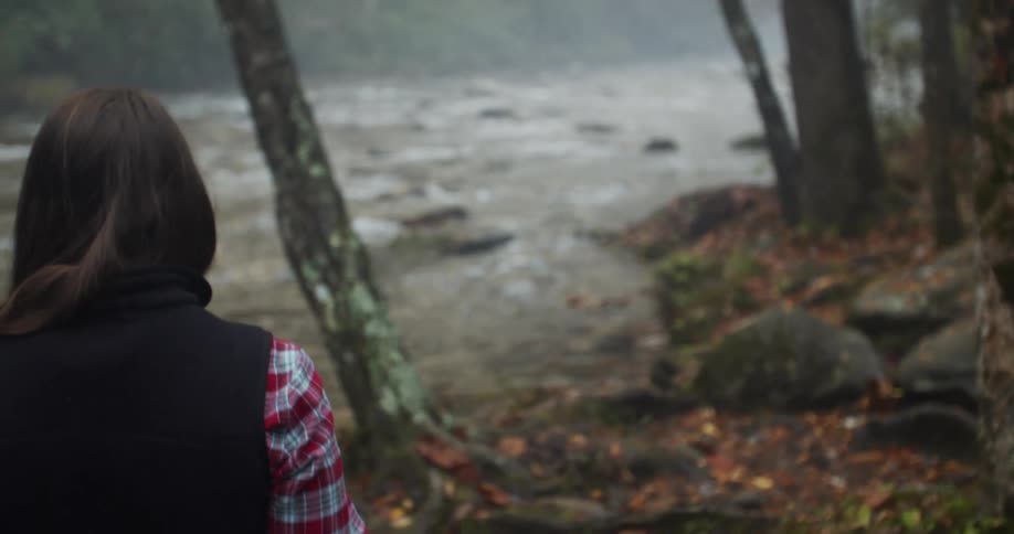 Woman wearing vest walks carefully over fallen leaves near flowing river