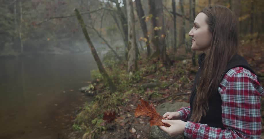 A woman standing and holding a leaf with a stream trees the sky and clouds in the background