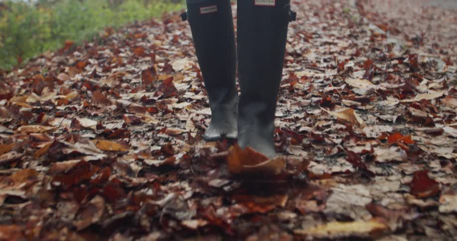A person in black rain boots walks through fallen leaves on the ground