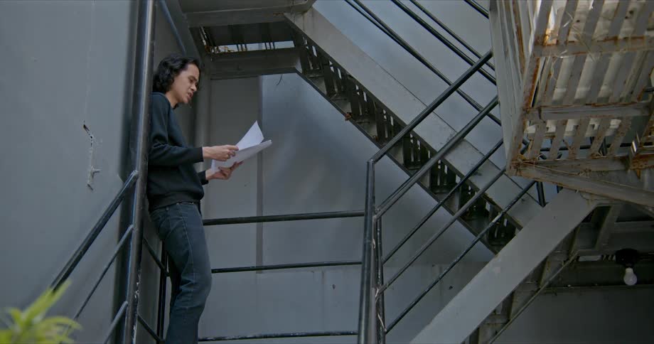 Man leans against wall on metal staircase landing reading papers