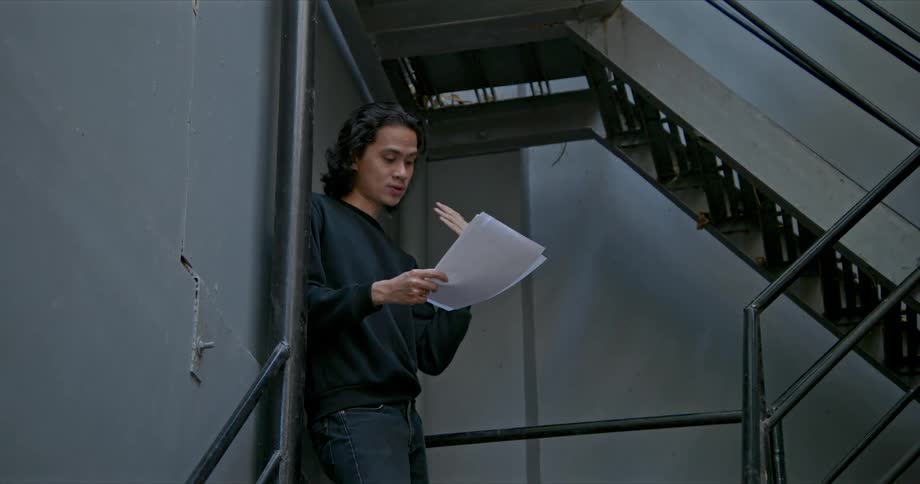 A teenager gestures while reading from sheets of paper while standing in a stairwell