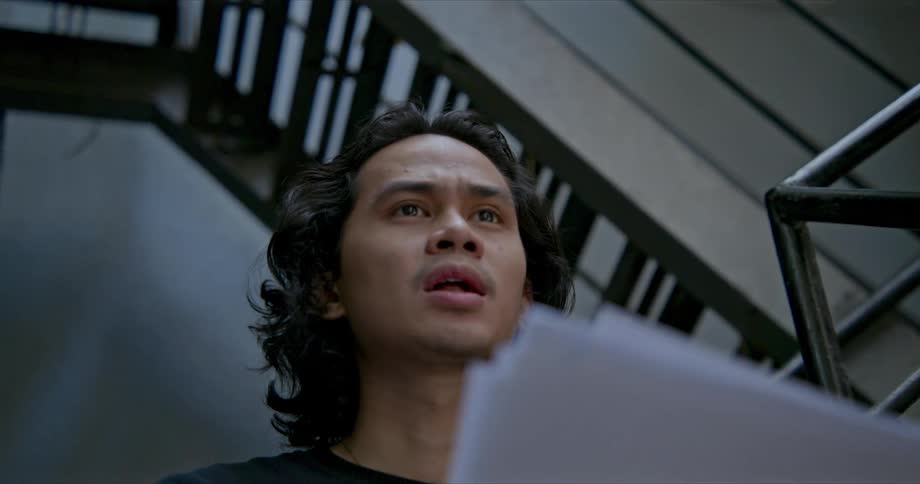 A man in a stairwell looks up from the script pages he is holding and says a line in an expressive manner
