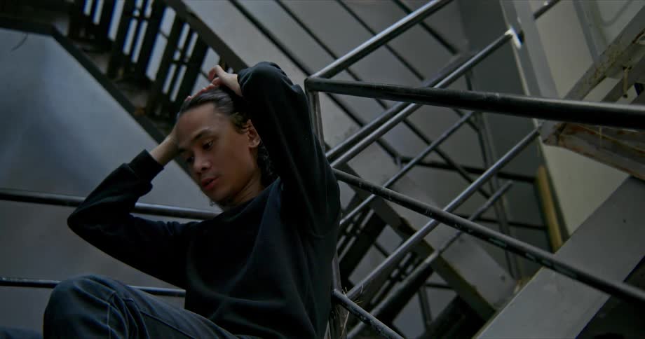 A young man sits in a stairwell looking sad and pulls back his hair