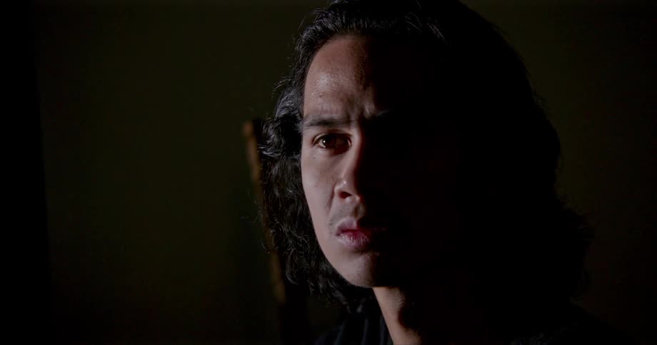 Man with brunette shoulder length hair focuses intently in shadowy room