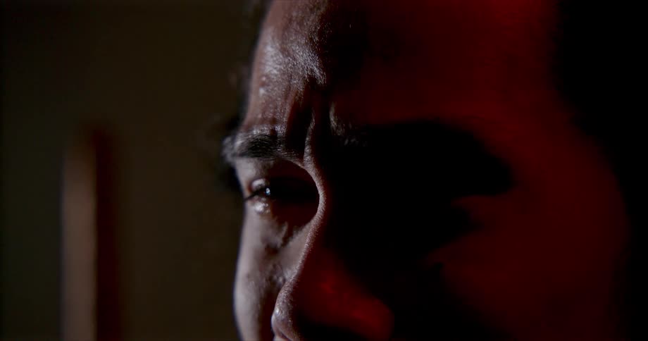 A man standing in the shadows grimaces as he cries and is sad
