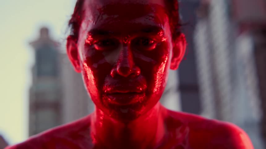Close up of a shirtless man covered in red paint under red lights in front of the city skyline