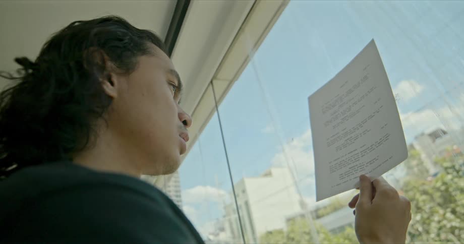 A man stands at a window holding up a sheet of paper reading it and studying