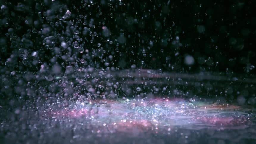 Drops of water landing on a surface just to be propelled into the air again with colorful lights flashing from above