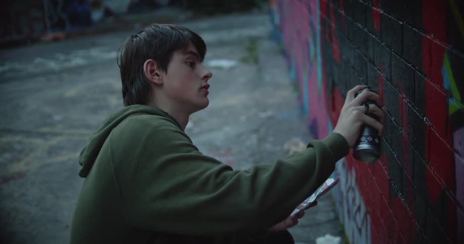 A young man spray paints a mural on a brick wall while holding a sketch of the design