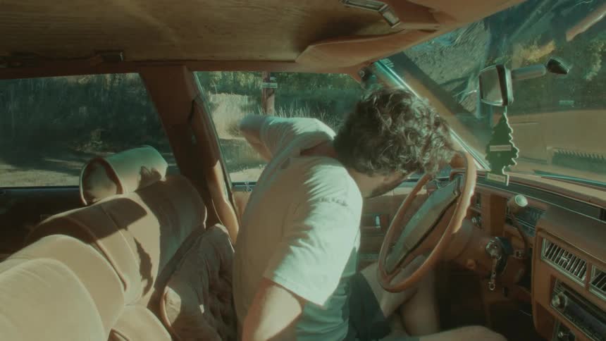 Man wearing shorts gets into car then sits with hand propped on door looking out front windshield