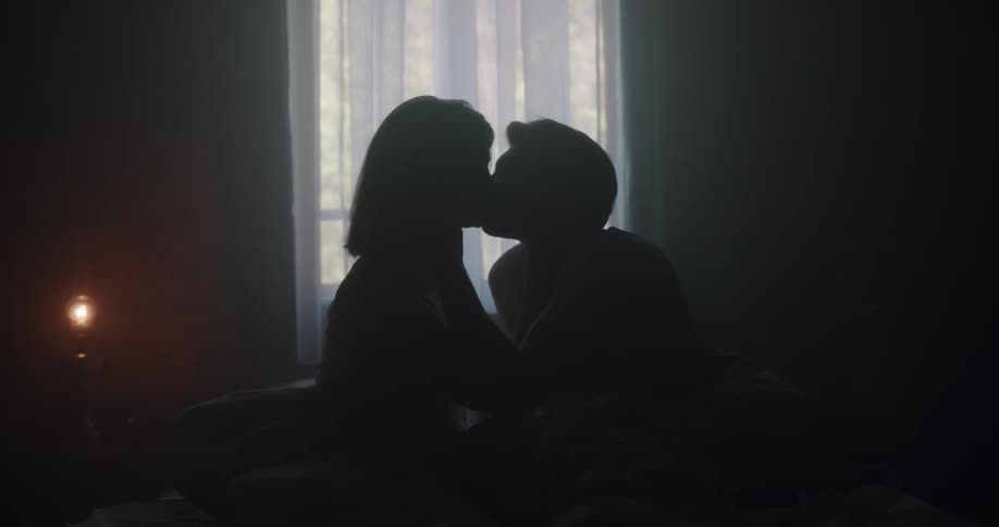 Couple sit on bed together kissing silhouetted by light from window in dark room