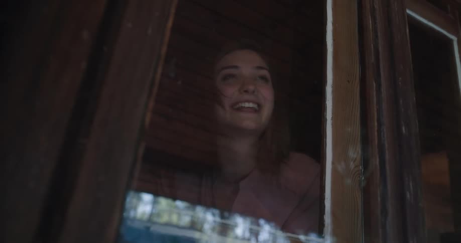 A young woman standing before a window smiles and waves before laughing a walking way