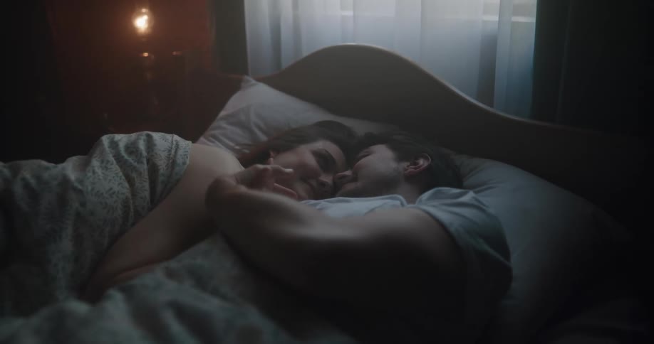 A man and a woman in a bedroom laying on a bed with a wall and window in the background