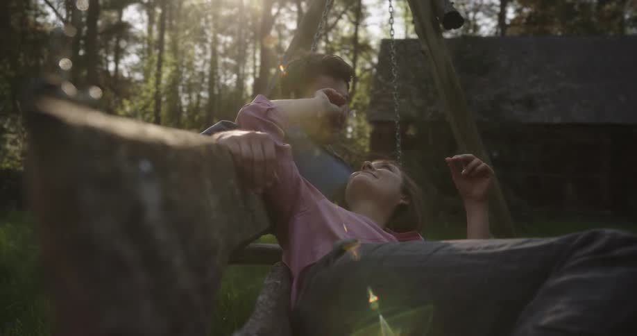 A man and a woman sitting on a swing with a cabin trees the sky and clouds in the background