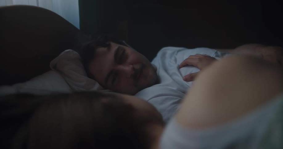 A woman runs her hand along her husbands chest as they lie in bed