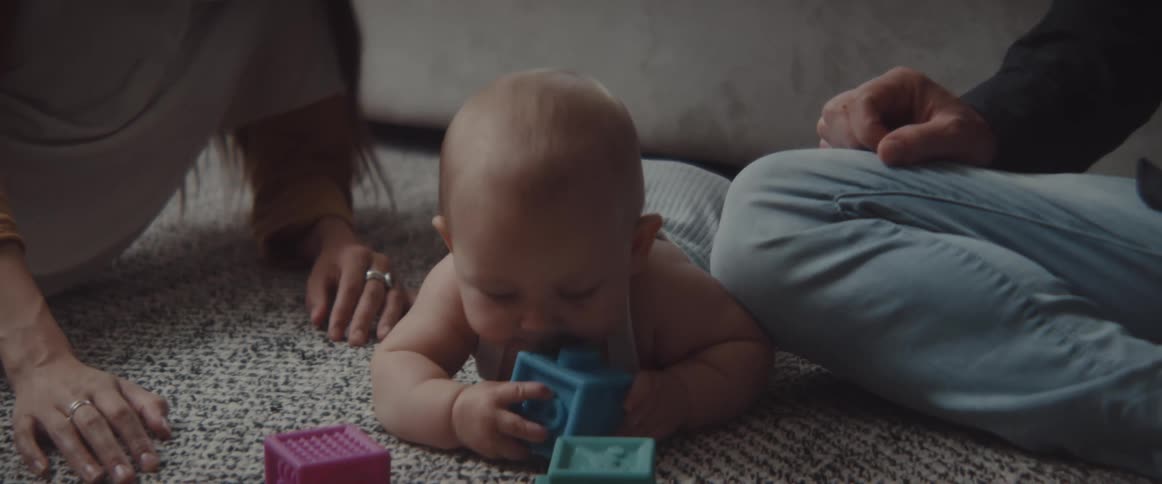 A baby lays on the floor chewing on toy blocks with two adults sitting next to them