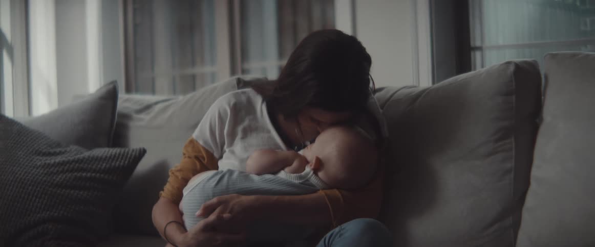 A woman sits on a couch holding a baby and kisses it on the head