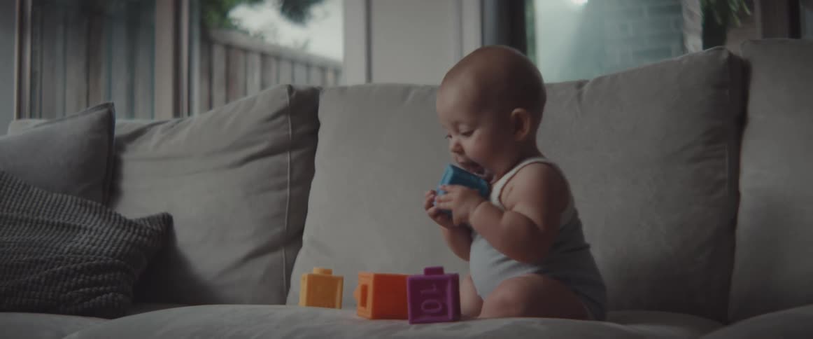 Baby plays with plastic blocks on couch set near window showing view of fence and tree