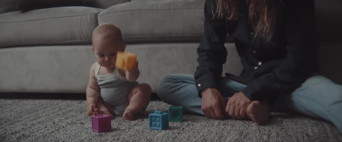 A baby plays with toy blocks as a man strokes their back while sitting on the floor in front of a sofa