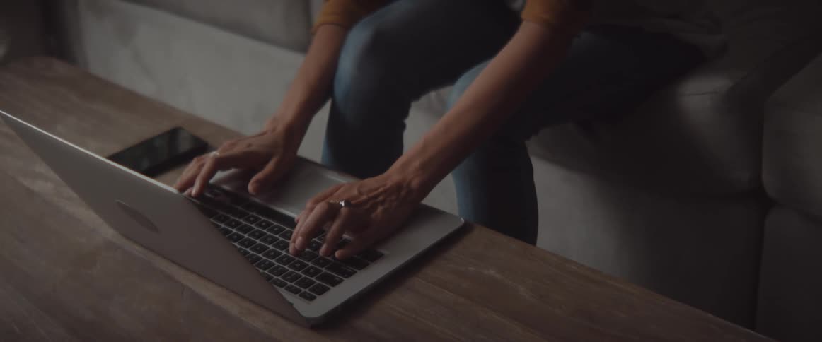 A woman in a room sitting on a couch typing on a laptop on a table
