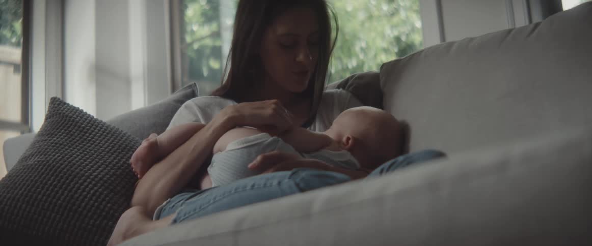 Mother cuddles and kisses baby sitting with legs under her on couch near windows showing trees outside