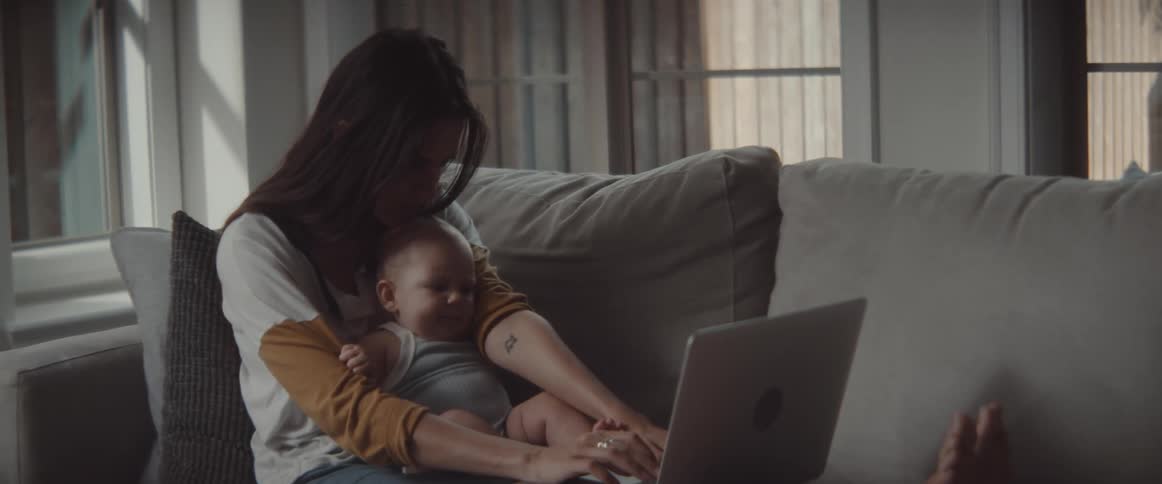 A woman in a room sitting on a couch holding a baby and using a laptop