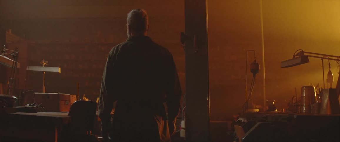 A graying man stands contemplatively in a dark office illuminated by orange sunlight from a window