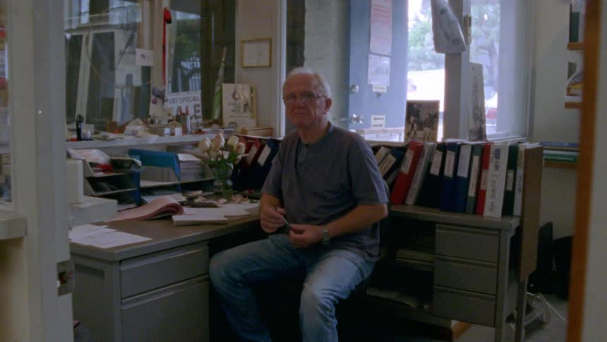 A man with glasses in a room sitting at a desk with a door in the background