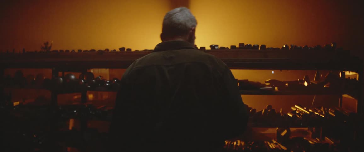 White haired man stands next to shelves full of items in shadowy room lit by amber light from across the room