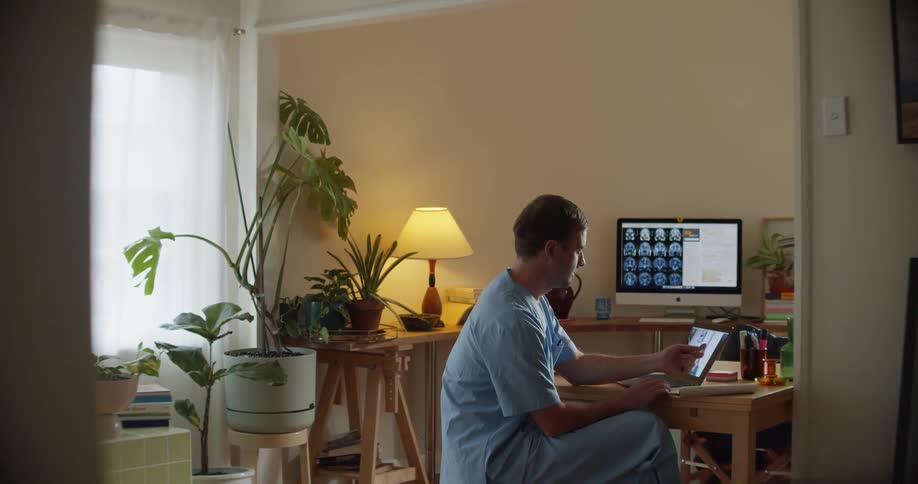 A nurse sitting at a table writes in a notebook while participating in a video chat on a laptop