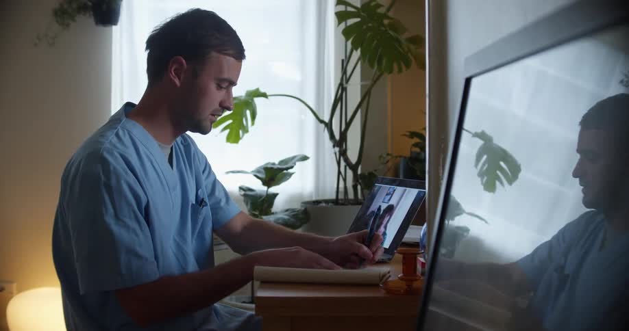 Man with a blue scrubs makes a video call on laptop and takes notes