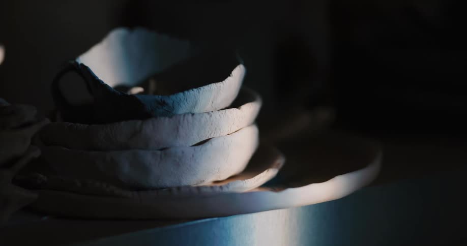 Stack of clay plates and bowl siton edge of metal platform in beam of light that is briefly obscured by passing shadow