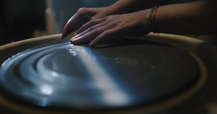 Woman turning a piece of clay on a pottery wheel inside a dimly lit room