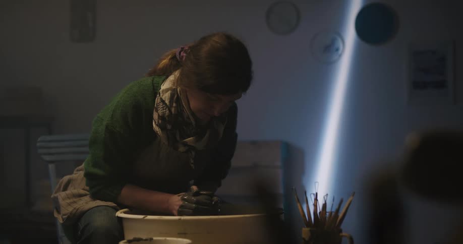 Woman works at pottery wheel her hands coated with clay in dimly lit workshop