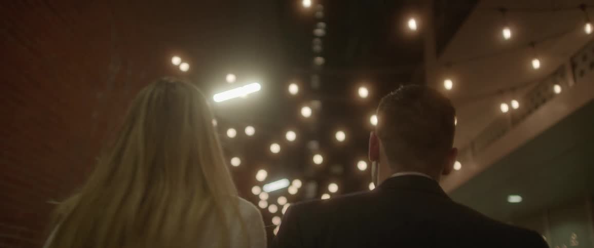 A man and a woman walking in an alley with buildings and lights in the background