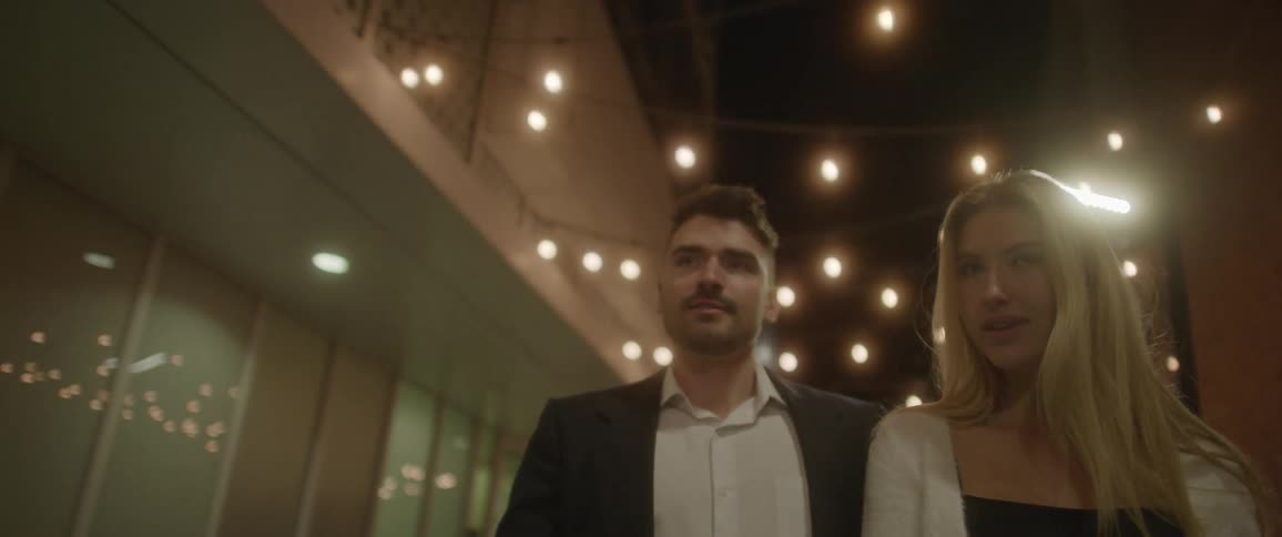 A man and woman walk through a city at night together with christmas lights strung on the buildings