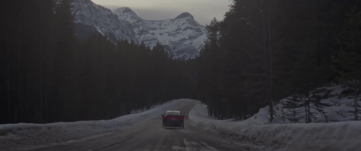 A black car travels on a snow covered road with trees along the roadside and snow covered mountains in the distance