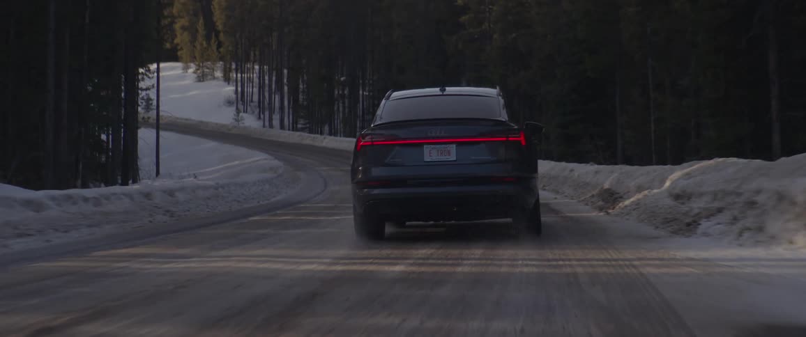 A sedan drives down a curvy snow covered road surrounded by tall trees