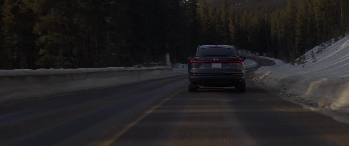 A black car travels on a road lined with tall trees and snow covered mountains in the distance
