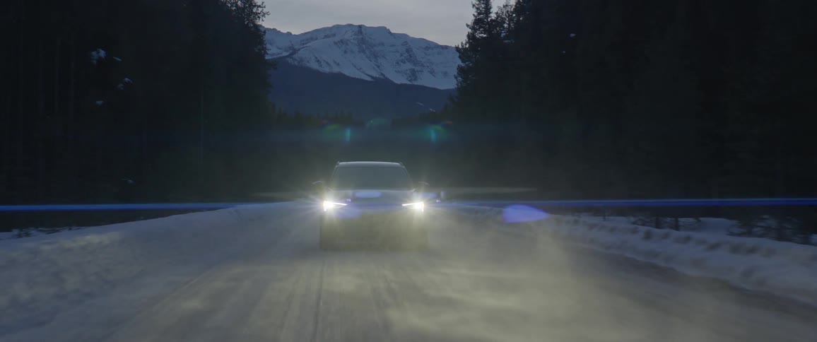 A black car travels on a snowy road with headlights on and a snow covered mountain in the distance