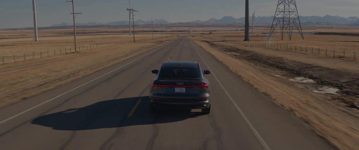 A black car travels on a road with wind turbines and a utility tower along the roadside