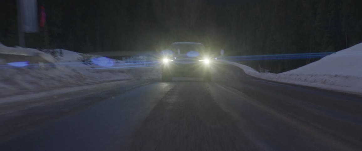 A black car with headlights on travels on a snow covered road with trees along the roadside