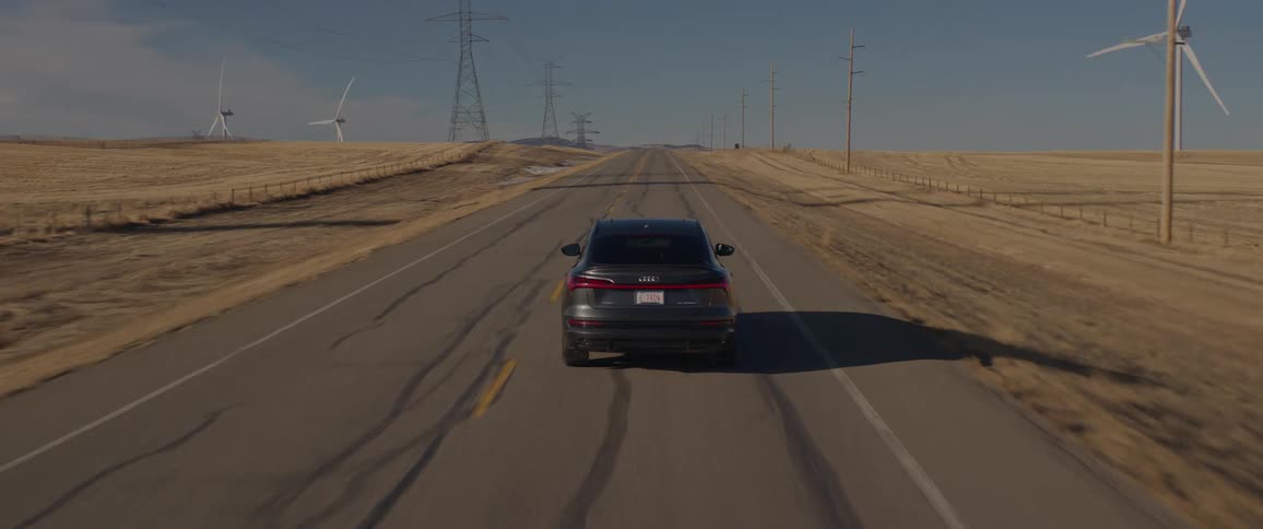 A car travels on a road with spinning wind turbines and utility towers along the roadside