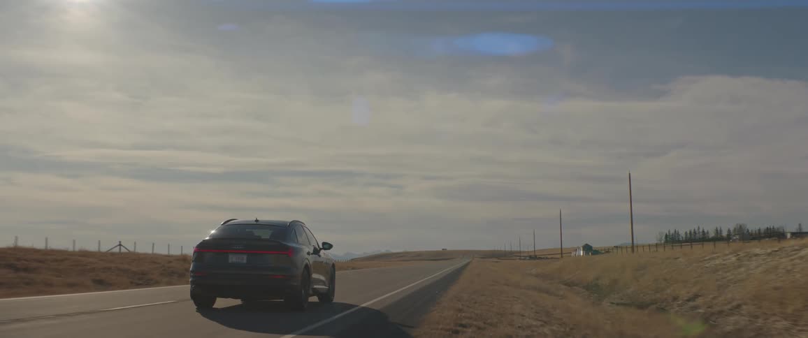 A vehicle drives down a rural highway past vast fields and an isolated farm house