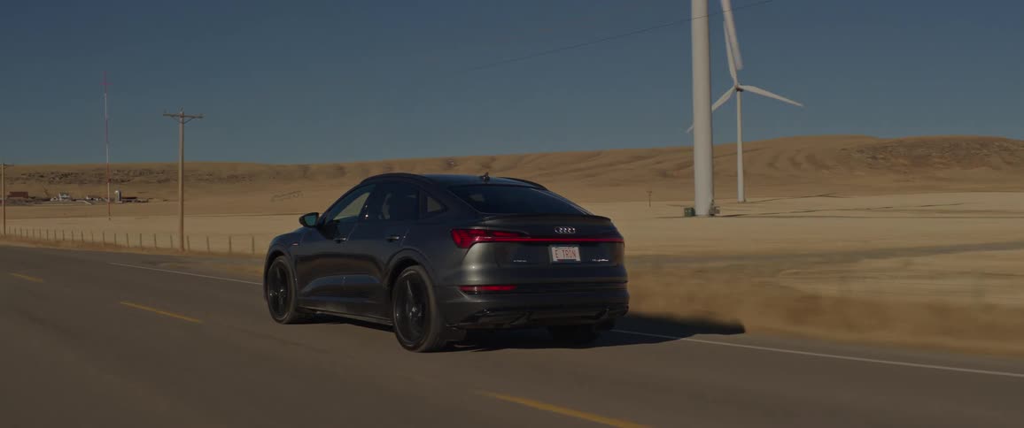 A black car travels on a road with spinning wind turbines and utility poles along the roadside