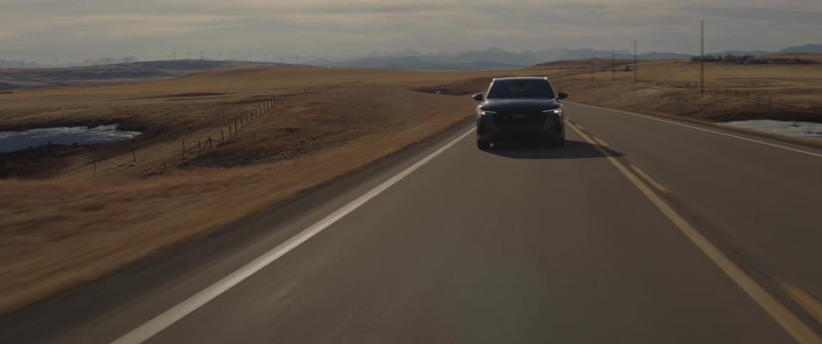 A black car travels on a two lane road with utility poles along the roadside