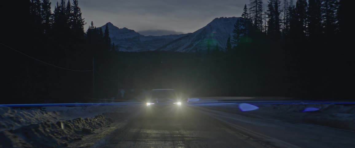 A black car with headlights on travels on a road lined with trees and snow covered mountains in the distance